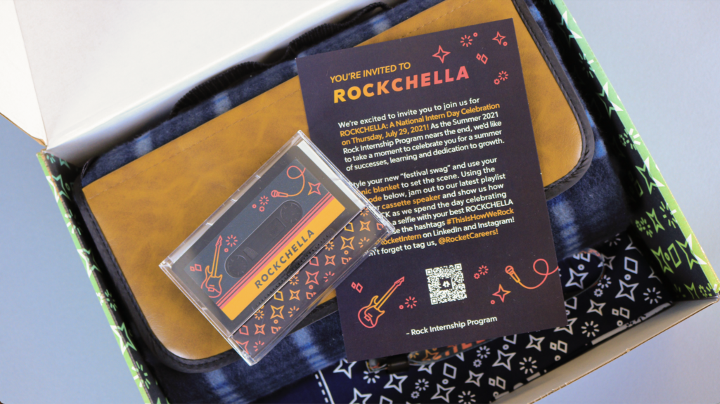 photo of the cassette tape and postcard inside of the Rockchella themed box that interns received them in. The cassette tape is inside of its plastic casing, with a guitar and microphone and the word "rockchella" on it. Behind it is a postcard that contains the same illustrations and the headline "you're invited to rockchella" accompanied by body copy that explains the event.