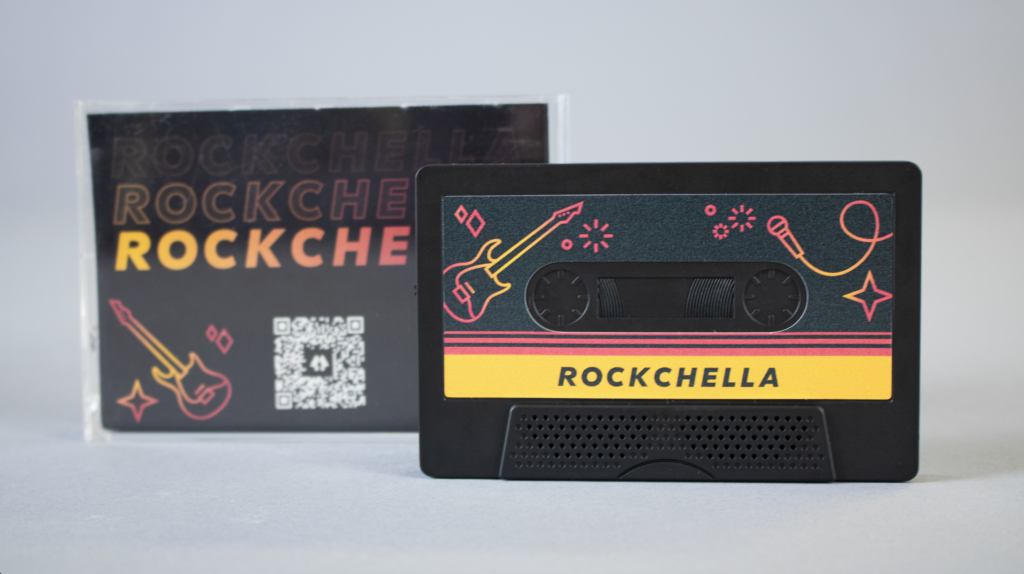 photo of the cassette tape outside of its plastic case, with the plastic case sitting behind it. The plastic case contains an illustration of a guitar, a QR code, and the text "rockchella." The cassette tape contains the same illustration of a guitar, as well as a microphone and the text "rockchella."