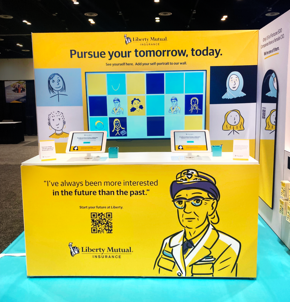 Photo of the empty Liberty Mutual Booth at the Grace Hopper conference. An illustration of Grace Hopper and a quote are on the front of a yellow stand, which is topped with touchscreens. Behind the stand is a wall with the text "pursue your tomorrow, today," and a digital display of a variety of doodled portraits on top of different colored squares.