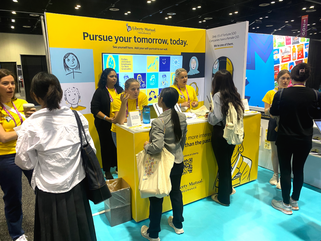 Photo of the Liberty Mutual Booth surrounded by people at the Grace Hopper conference. An illustration of Grace Hopper and a quote are on the front of a yellow stand, which is topped with touchscreens. Behind the stand is a wall with the text "pursue your tomorrow, today," and a digital display of a variety of doodled portraits on top of different colored squares.