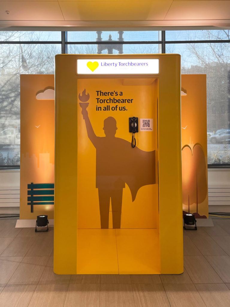 Photo of a yellow phone booth. At the top is a glowing sign that says, "Liberty Torchbearers." Inside is a phone, a QR code, and the silhouette of a person with a cape holding a torch high. Above the silhouette is the text, "There's a Torchbearer in all of us."