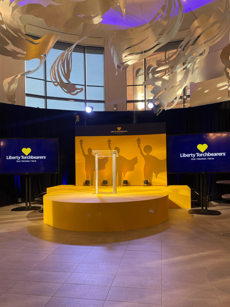 Photo of a yellow stage in a corporate building. Digital displays on either side of the stage display the Liberty Torchbearers logo on a dark blue background. The stage hosts a podium, and a backing panel of three silhouettes of people wearing capes and holding torches high printed on a yellow background.
