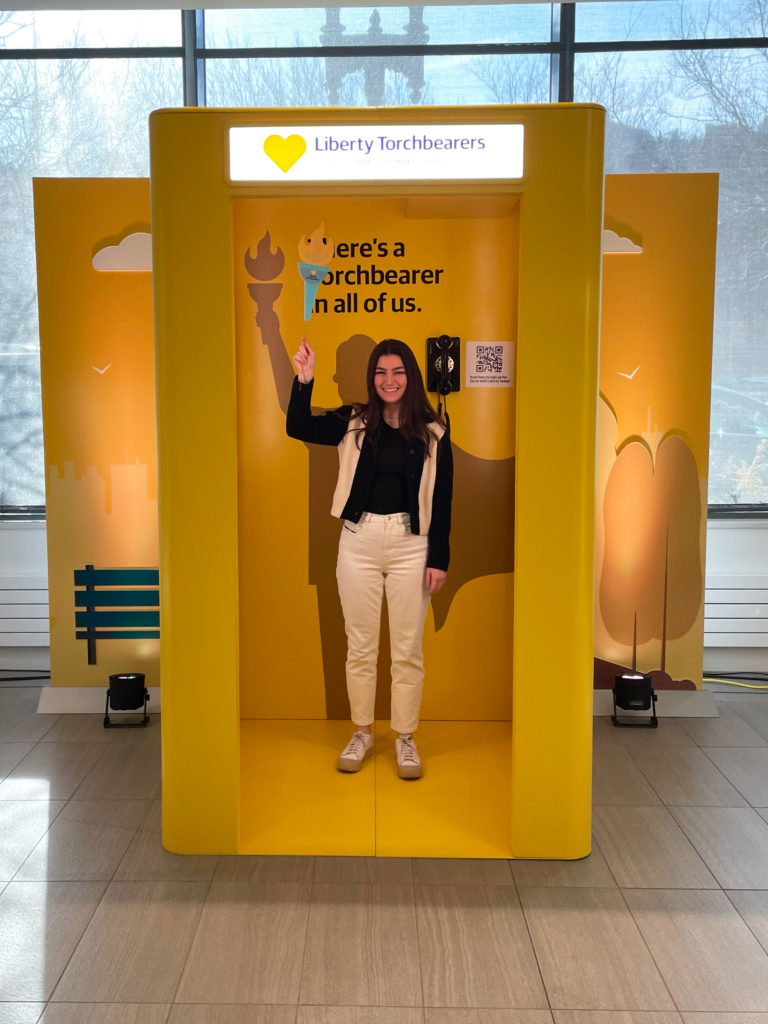 Photo of a yellow phone booth. A person stands inside, smiling and holding up a prop torch. At the top is a glowing sign that says, "Liberty Torchbearers." Inside is a phone, a QR code, and the silhouette of a person with a cape holding a torch high. Above the silhouette is the text, "There's a Torchbearer in all of us."