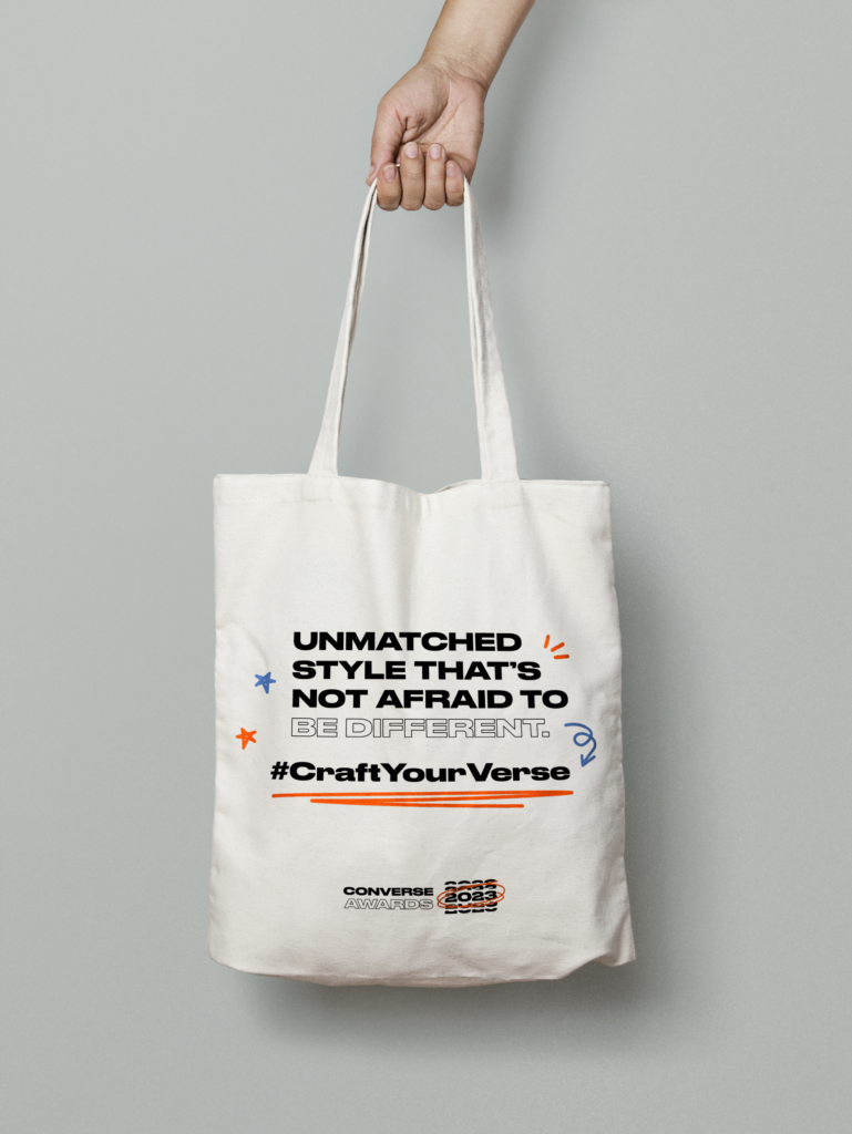 Mockup of a hand holding a white tote bag. On the tote bag is text that reads, "Unmatched style that's not afraid to be different. #CraftYourVerse."