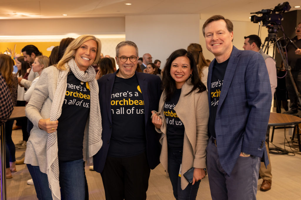 Photo of four people smiling for the camera. They are each wearing a dark blue shirt with yellow and white text on it that reads, "There's a Torchbearer in all of us."