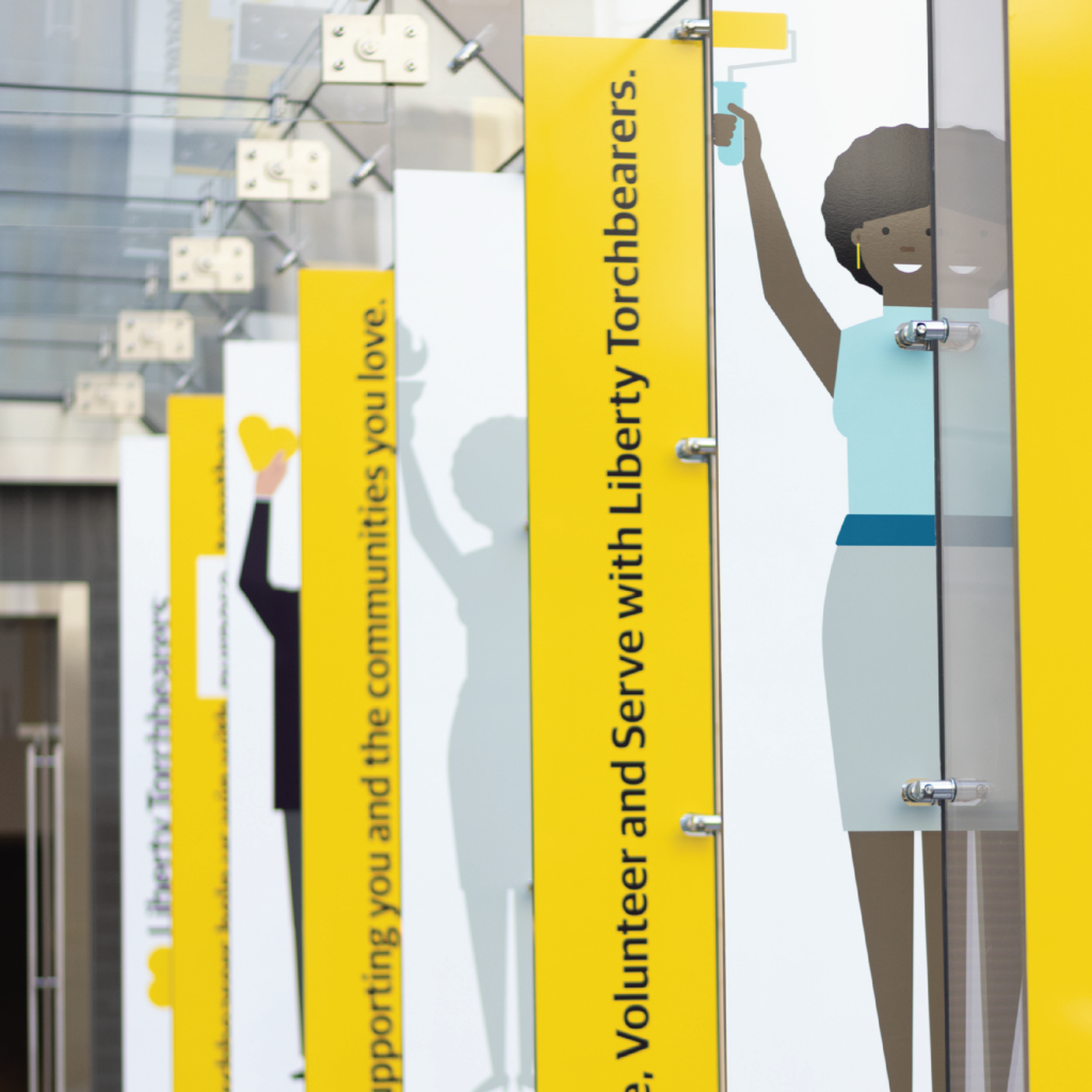Photo of several banners affixed to a glass wall. one after another. They contain corporate illustrations of people holding up a paint roller and a heart, as well as the text "volunteer and serve with liberty torchbearers."