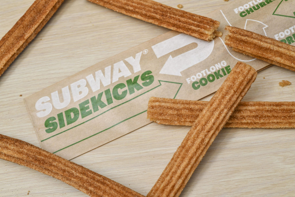 Overhead-angle photo of several footlong churros surrounding a long, thin sleeve of brown paper that displays the text "Subway Sidekicks," "Footlong cookie," "footlong churro," and "footlong pretzel" in green and white ink.