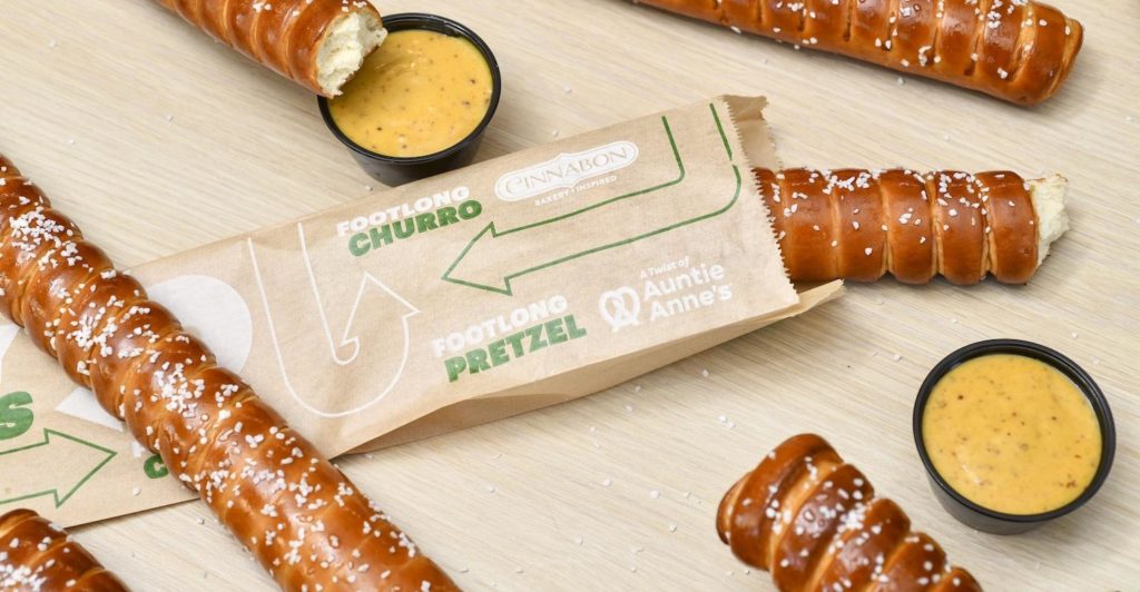 Overhead-angle photo of several footlong pretzels surrounding a long, thin sleeve of brown paper that displays the text "footlong churro," and "footlong pretzel" in green and white ink.