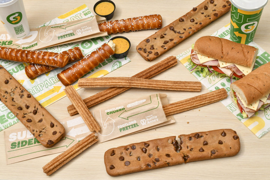 Overhead-angle photo of several footlong churros, cookies, and pretzels, as well as subway drinks and sandwiches surrounding a long, thin sleeve of brown paper that displays the text "Subway Sidekicks," "Footlong cookie," "footlong churro," and "footlong pretzel" in green and white ink.