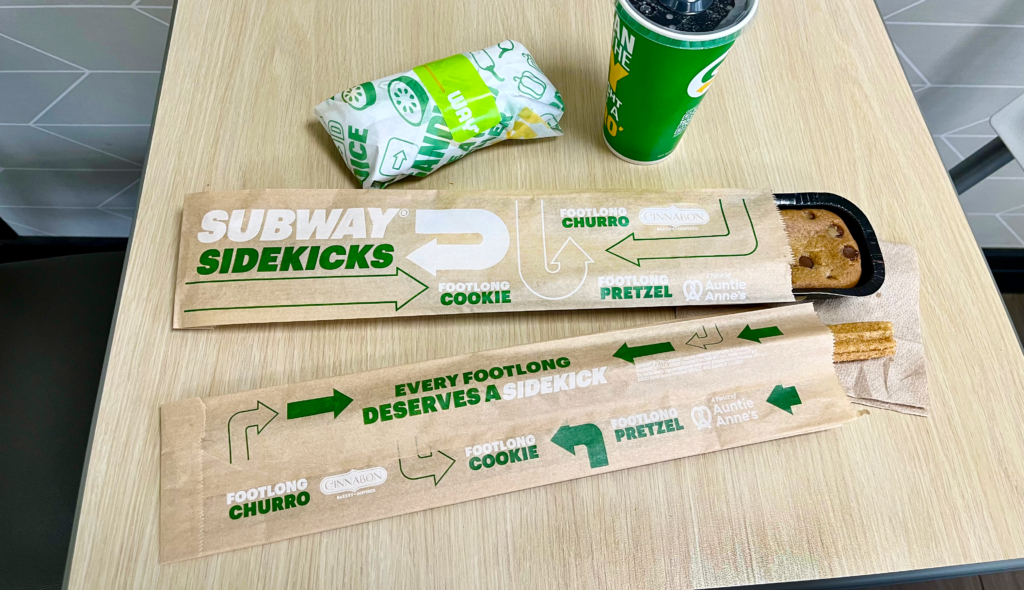 Overhead-angle photo of a footlong cookie and footlong churro emerging from two long, thin sleeves of brown paper that display the text "Subway Sidekicks," "Footlong cookie," "footlong churro," "footlong pretzel," the phrase "every footlong deserves a sidekick," and the Subway, Auntie Anne's, and Cinnabon logos in green and white ink.