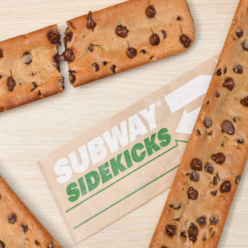 Overhead-angle photo of several footlong Subway cookies surrounding a long, thin sleeve of brown paper that displays the text "Subway Sidekicks" in green and white ink.
