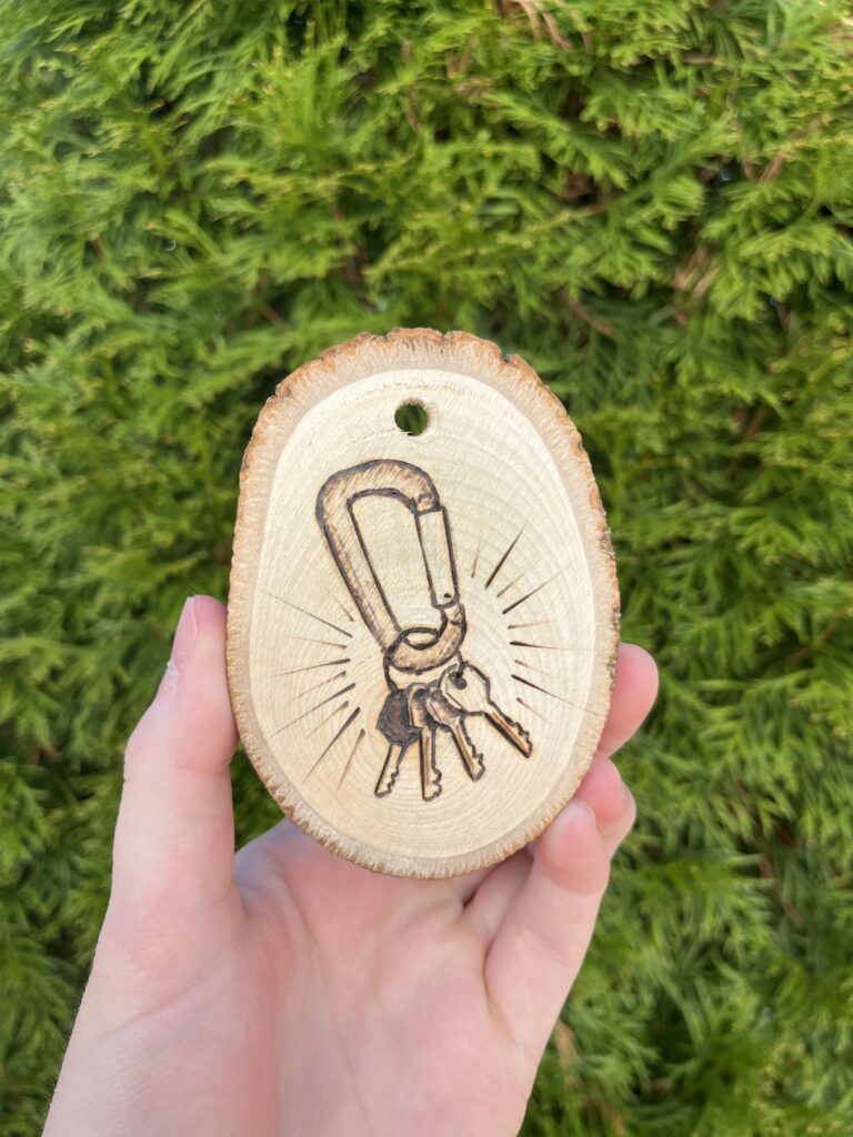 Photo of a hand holding a small wooden disc that is a cross-cut of a tree. A carabiner with keys on it is burned into it.