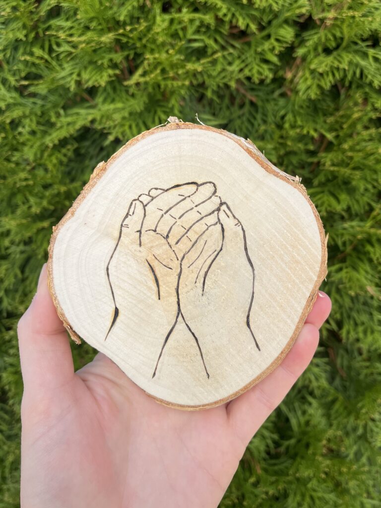 Photo of a hand holding a small wooden disc that is a cross-cut of a tree. An illustration of two cupped hands is burned into it.