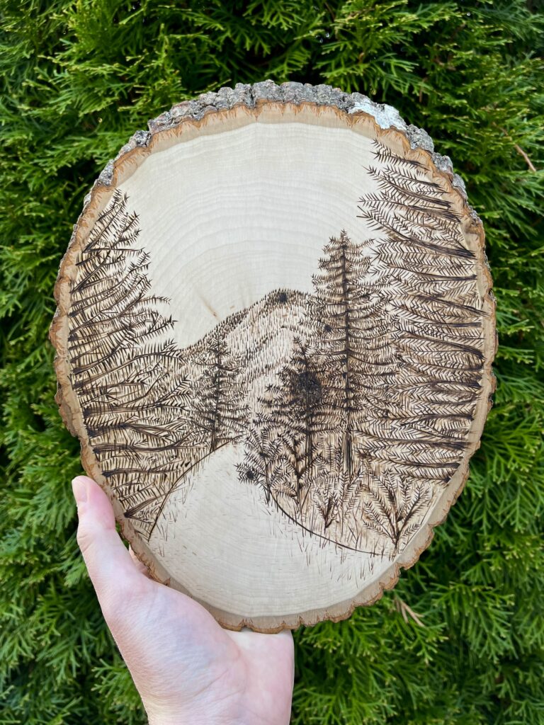 Photo of a hand holding a large wooden disc that is a cross-cut of a tree. An illustration of a mountain is burned into it.